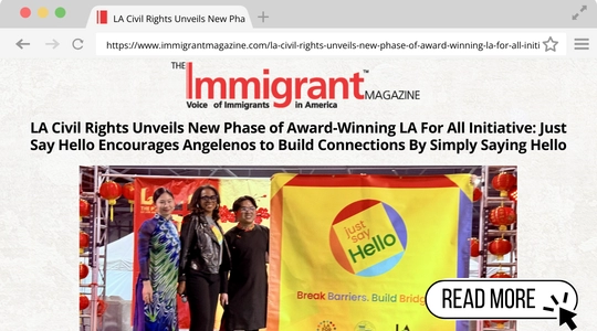 Screenshot of an article from The Immigrant Magazine titled 'LA Civil Rights Unveils New Phase of Award-Winning LA For All Initiative: Just Say Hello Encourages Angelenos to Build Connections By Simply Saying Hello.' The photo shows four people standing in front of a colorful 'Just Say Hello' banner with the tagline 'Break Barriers. Build Bridges.' The event setup includes festive red lantern decorations."