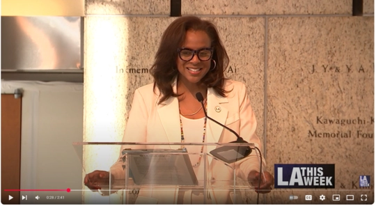 Video image from LA This Week of Capri Maddox at podium in front of memorial wall introducing panel for AANHPI Discussion