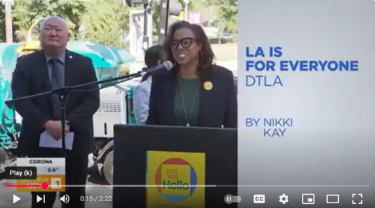 Capri Maddox speaks at a podium launching the new 'Just Say Hello' campaign sign, while a man stands nearby. On-screen text reads 'LA IS FOR EVERYONE DTLA by Nikki Kay.