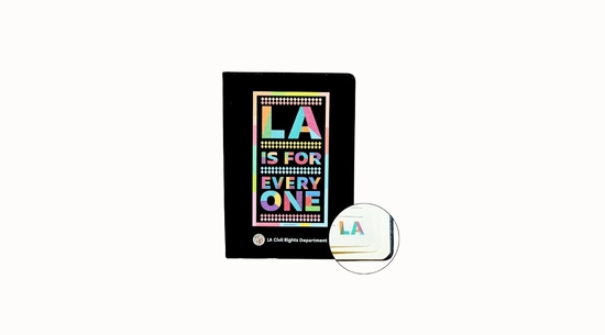 A closed, black journal with a multi-colored logo that says "LA IS FOR EVERY ONE." The journal has a matching  "LA" logo on the bottom right of each ruled page.
