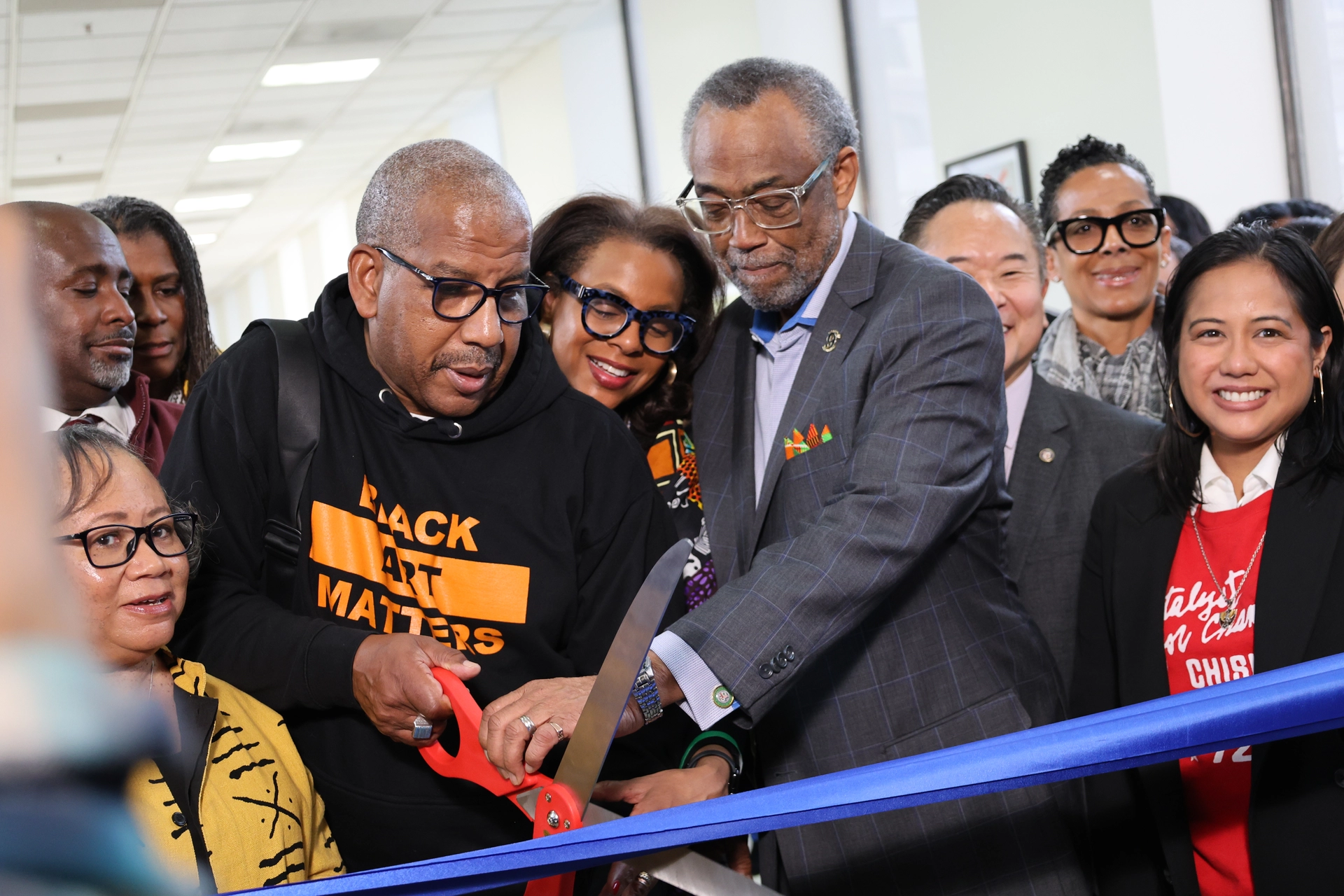 Image shows crowd Councilmember Curren Price holds scissors with Leroy Hamilton and Capri Maddox.