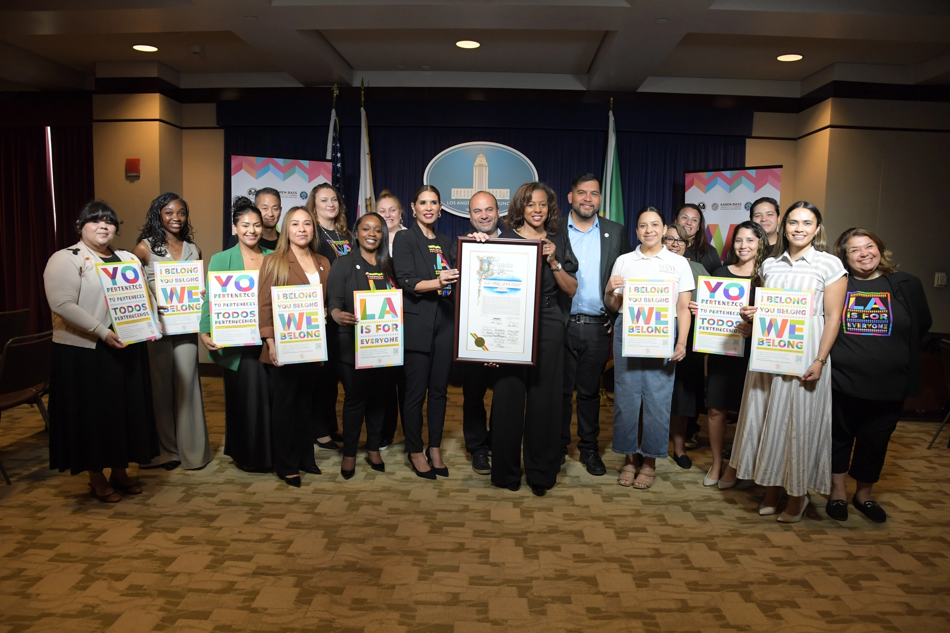 City leaders and community partners stand together in a formal room for a group photo to launch the 4th annual LA For All campaign. Officials in the center hold a framed proclamation, while others hold colorful signs that read, "I Belong. You Belong. We Belong." and "Yo Pertenezco. Tú Perteneces. Todos Pertenecemos."