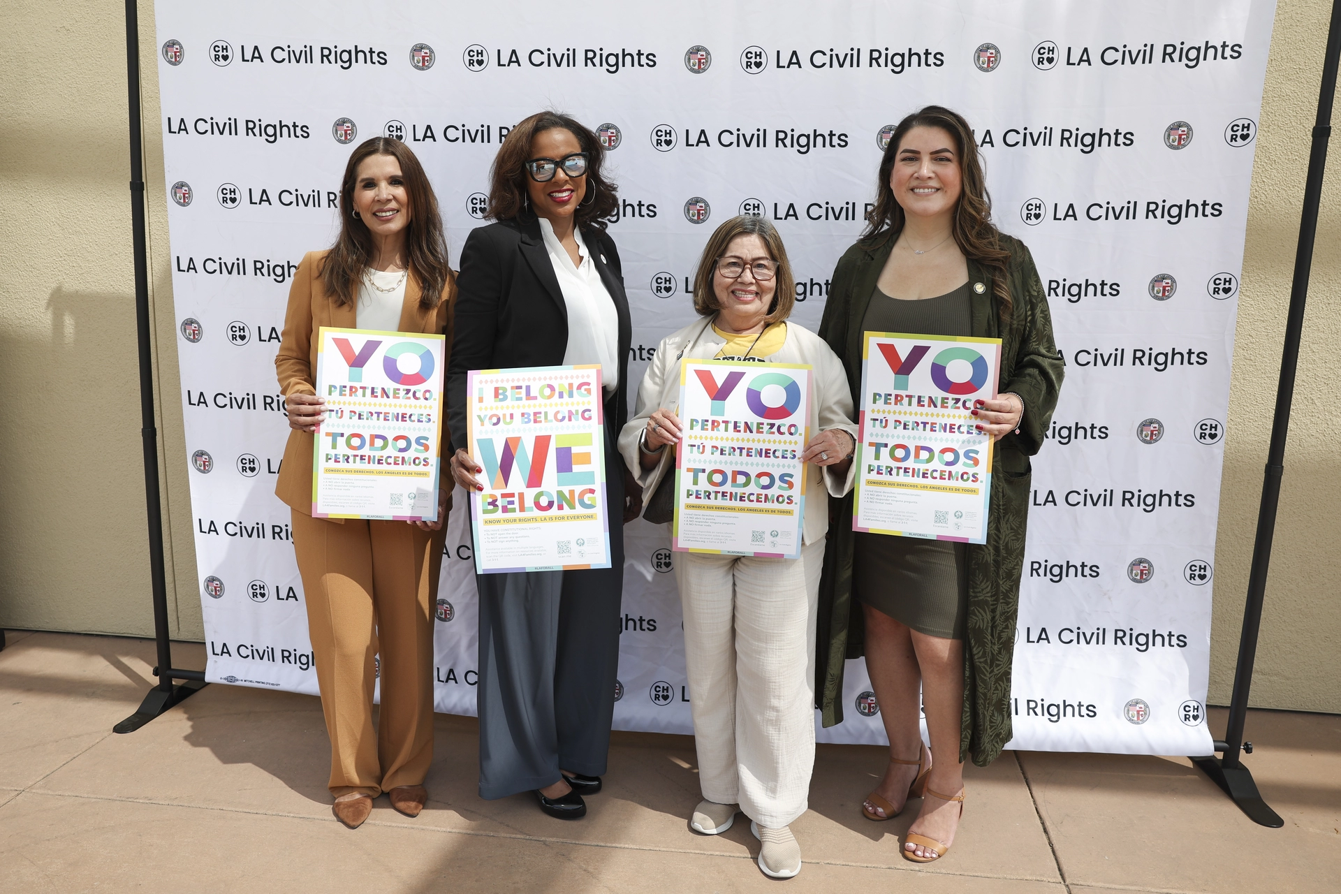 Four women pose holding We Belong signs in English and Spanish from Left to Right: Abigail Marquez, CIFD; Capri Maddox, LA Civil Rights, Dr Rosa Heckenberg, Cal State LA Dreamers Resource Center, and Claudia Aragon, Office of the Mayor of Los Angeles.