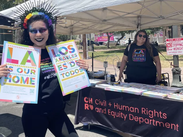 A young woman holds two LA For All posters in English and Spanish in front of the LA Civil Rights table at Boyle Heights Pride.