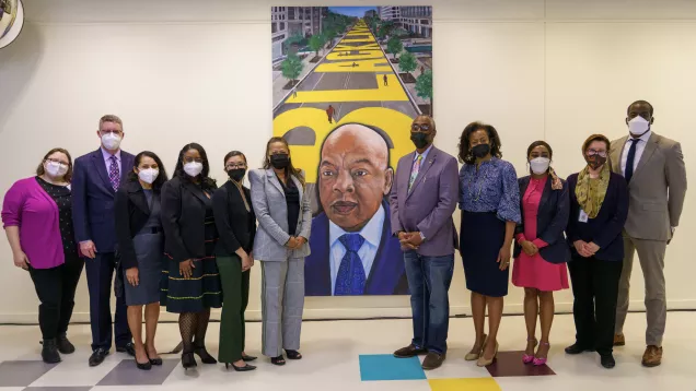 11 individuals stand before a large portrait of Congressmember John Lewis.