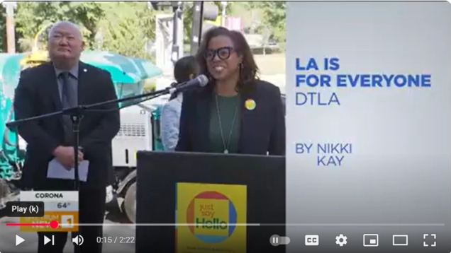 Capri Maddox speaks at a podium launching the new 'Just Say Hello' campaign sign, while a man stands nearby. On-screen text reads 'LA IS FOR EVERYONE DTLA by Nikki Kay.